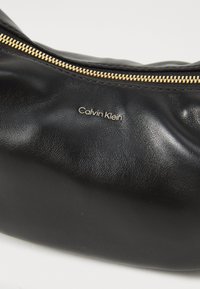 Geantă de mână din piele neagră cu un fermoar auriu și logo-ul „Calvin Klein” embosat. Textură moale, formă rotunjită și design minimalist.