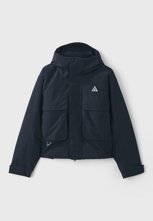 Veste bleu marine avec capuche, deux grandes poches avant et un logo triangulaire. Texture lisse avec fermeture éclair et poignets ajustables.