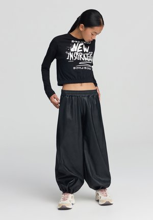 Maglietta nera a maniche lunghe e a cropped con testo grafico bianco, abbinata a pantaloni neri larghi. L'outfit presenta un elastico in vita e un materiale testurizzato.