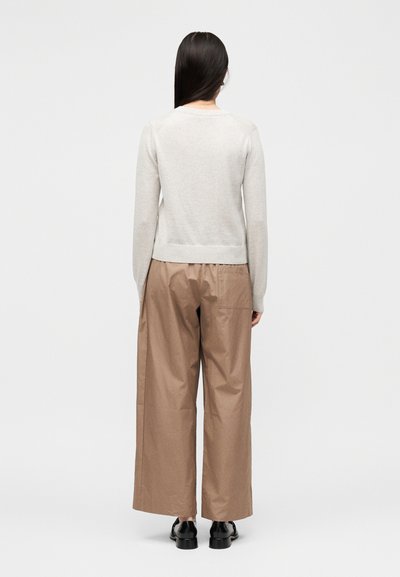 Γυναίκα με μακριά σκούρα μαλλιά στέκεται με την πλάτη, φορώντας ένα ανοιχτό γκρι πουλόβερ, άνετο ανοιχτό καφέ παντελόνι και μαύρα παπούτσια σε απλό φόντο.