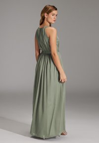 Lange, mouwloze groene jurk met een aansluitende taille en luchtige rok, met een geplooide achterkant en subtiele glans. Zilveren sandalen met hakken zichtbaar.