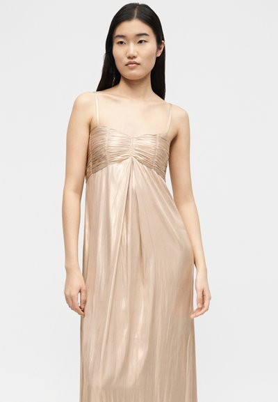 Νεαρή γυναίκα με μακριά σκούρα μαλλιά, ντυμένη με λαμπερό, ανοιχτό μπεζ, αμάνικο φόρεμα με λεπτές τιράντες και ρυτιδωμένο μπούστο.