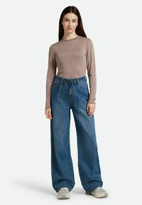 Top de manga larga color marrón claro, ajustado al cuerpo. Pantalones vaqueros de denim azul de pierna ancha con un ajuste ligeramente holgado y cintura con cordón. Zapatillas neutrales.