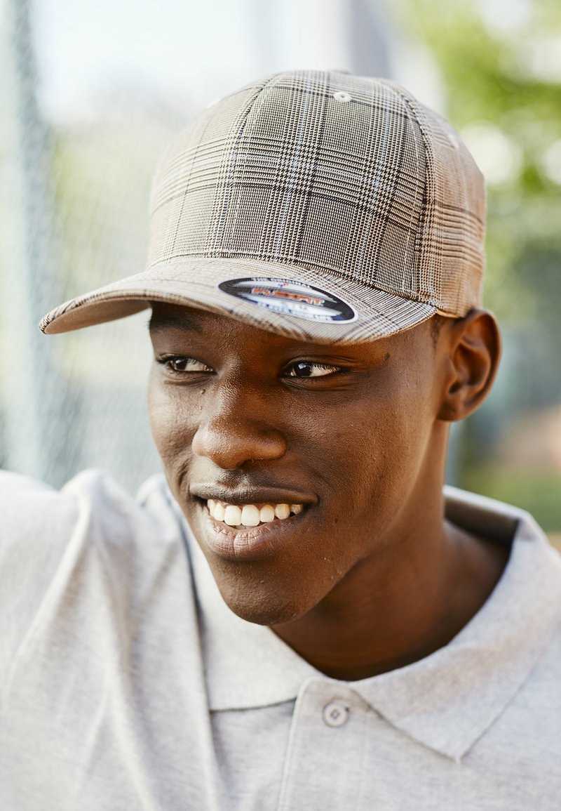 Baseball cap med et lyst beige, ternet mønster, forsynet med en buet skygge og et fremtrædende brandmærke foran. Båret med en grå polo shirt.