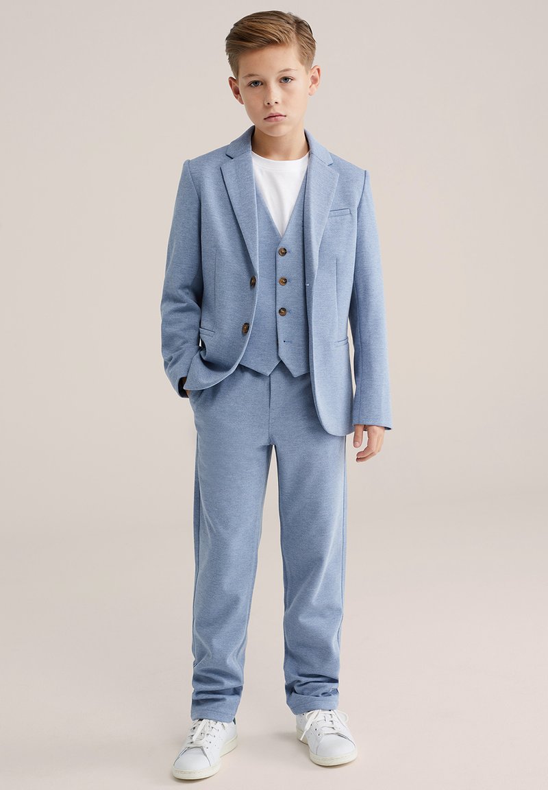 Világoskék öltöny, testhezálló zakóval és hozzáillő nadrággal. Az anyag texturált és sima, háromgombos mellénnyel és fehér pólóval.