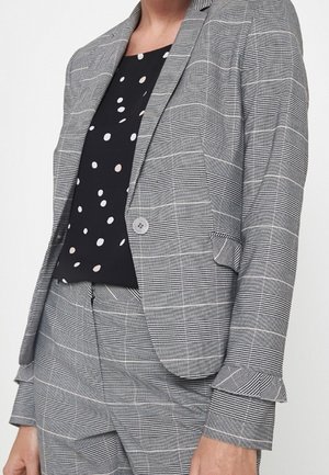 Femme portant un blazer à carreaux gris et un pantalon assorti, avec un chemisier noir orné de pois blancs et beiges.