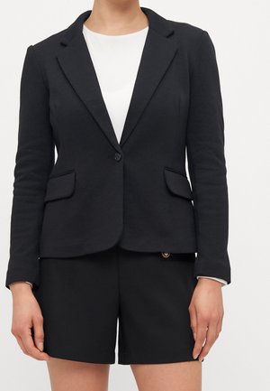 Frau trägt einen schwarzen taillierten Blazer mit einem Knopf und Pattentaschen, kombiniert mit schwarzen Shorts und einem weißen Top darunter.