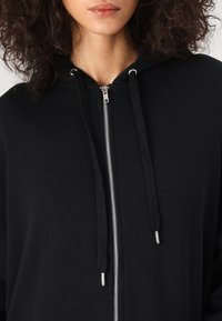 Svart zip-up hoodie i mjukt tyg, med en silverfärgad dragkedja, snören och en avslappnad passform.