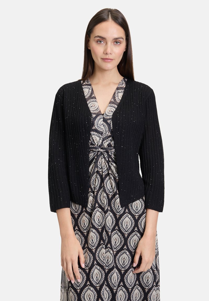 Cardigan noir à côtes avec des manches trois-quarts, orné de accents argentés dispersés. Porté sur une robe à motifs en V dans des tons terreux.