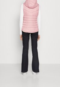 Ljusrosa pufferväst med huva, med ett quiltat mönster och utsvängda mörka jeans. Outfiten fullbordas med vita sneakers.