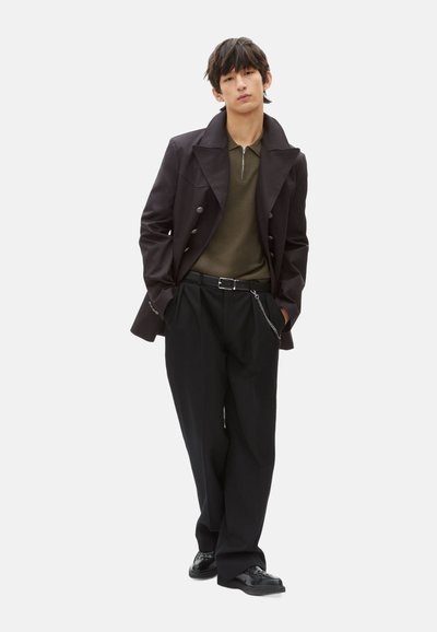Dunkler, doppelt geknöpfter Mantel mit großen Revers, olivgrünes Polo mit Reißverschluss, schwarze weite Hose und schwarze Anzugschuhe.