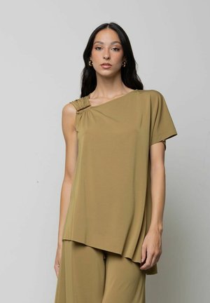 Donna con lunghi capelli neri che indossa orecchini a cerchio dorati e un top ampio asimmetrico verde oliva con pantaloni abbinati su sfondo uniforme.