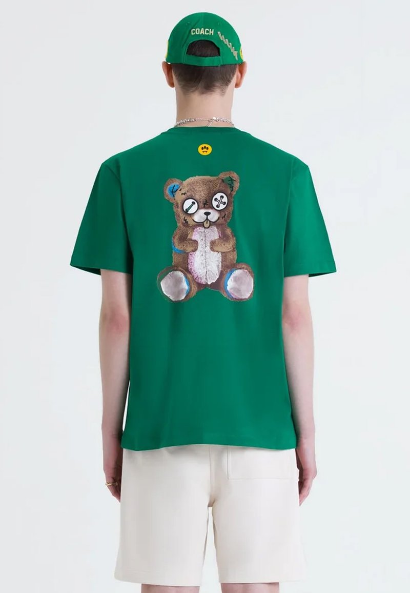 T-shirt in cotone verde con una grafica di un orso cartoon sulla schiena, con occhi rotondi e dettagli cuciti. Completato da pantaloni corti color crema.