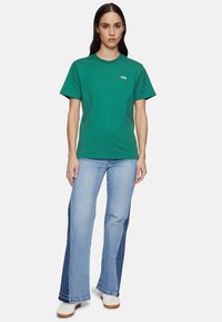 Groene katoenen t-shirt met korte mouwen en een ronde hals, met een witte FILA-logo; gecombineerd met uitlopende denim jeans en witte sneakers.