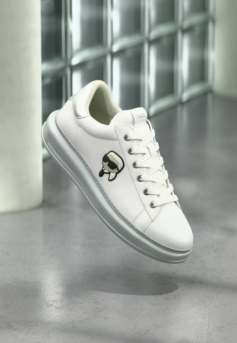 Λευκό αθλητικό παπούτσι με κορδόνια και μικρό καρτούν γραφικό στην πλευρά, παρουσιάζεται μπροστά σε μια ανακλαστική, πλακιδωτή επιφάνεια και γκρι πάτωμα.