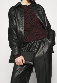 Černá kožená košile s bundou přes svetr v barvách burgundské a černé zebry, doplněná černými koženými kalhotami s vysokým pasem a psaníčkem.