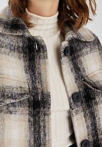 Kostkovaná bunda z vlněné směsi v béžové, černé a krémové barvě s velkými knoflíky a náprsními kapsami, vrstvená přes žebrovaný světle béžový top.