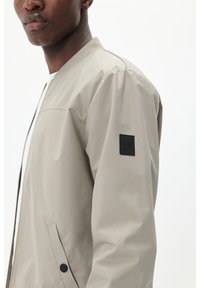 Beige Bomberjacke mit glatter Textur, geripptem Kragen, Reißverschluss und einem schwarzen rechteckigen Logo-Patch auf dem linken Ärmel.