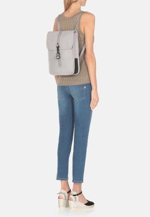 Sac à dos en cuir gris avec un rabat et des bretelles réglables, porté par une personne, portant un haut beige à côtes et un jean bleu.