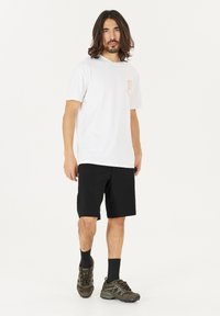 Man met schouderlang haar, gekleed in een wit T-shirt met opdruk, zwarte shorts, zwarte sokken en bruine wandelschoenen, staande tegen een witte achtergrond.