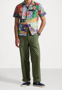 Uomo che indossa una camicia a maniche corte a fiori colorata in patchwork, pantaloni verdi con risvolto, sneakers in canvas blu navy e un orologio da polso, in piedi con una mano in tasca.