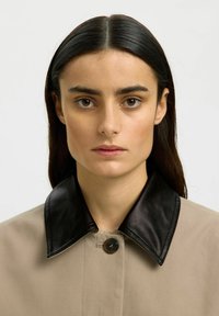 Femme aux longs cheveux foncés portant un manteau beige avec un grand col en cuir noir, face à l'avant sur un fond clair uni.