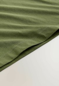 Olivgrüner Baumwollstoff drapiert, der eine glatte Textur und eine gefaltete Kante mit kontrastierender Naht zeigt. Leichter Kontrast auf der Oberfläche.