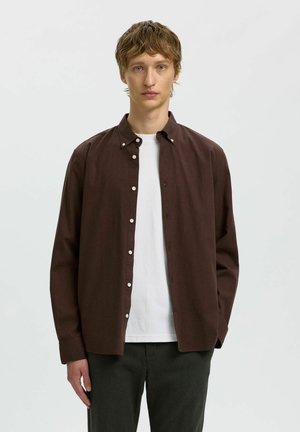 Jeune homme aux cheveux châtain clair portant une chemise marron foncé boutonnée par-dessus un t-shirt blanc et un pantalon sombre, debout devant un fond uni clair.