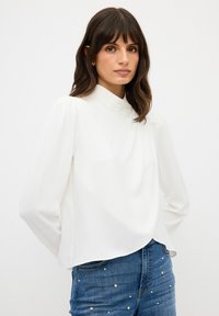 Blusa bianca a maniche lunghe con colletto alto, in tessuto morbido, con delicati plissè sulle spalle, abbinata a jeans in denim blu con applicazioni di perle.