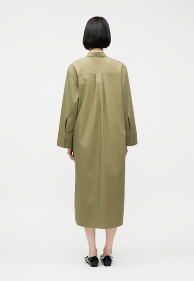 Άτομο με κοντά μαύρα μαλλιά, που φοράει ένα μακρύ, ελαιδί παλτό και μαύρα παπούτσια, στέκεται στραμμένο με την πλάτη σε απλό λευκό φόντο.