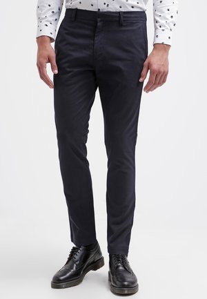 Hombre con pantalones ajustados azul marino oscuro, zapatos de vestir de cuero negro y una camisa blanca con lunares negros visibles en los puños y la cintura.