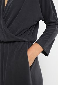 Combinaison noire à manches longues en tissu extensible, avec une coupe décontractée et un détail wrap. Offre des poches latérales et une texture subtile.