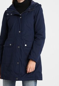 Námořnická zateplená bunda s předním zapínáním na zip a kovové knoflíky, nastavitelnou kapucí a dvěma předními kapsami se zaklapávací klopou.
