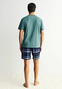 Lyhythempparinen teal-värinen puuvillapaita yhdistettynä tummansinisiin ruutuhousuihin, joissa on valkoisia ja sinisiä yksityiskohtia, esiteltynä takaa.