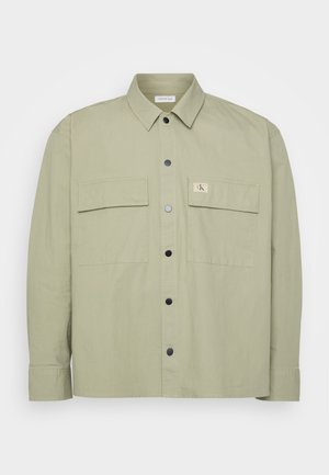 Lys grønn skjorte med knapper laget av mykt stoff. Den har to store brystlommer og et CK-logo-patch. Sorte trykknapper fullfører designet.