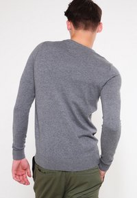 Šedý svetr s dlouhými rukávy a kulatým výstřihem, pouzdrového střihu, hladké textury, s vroubkovanými manžetami a lemem, nošený přes zelené kalhoty.