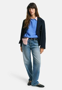 Blazer navy, felpa azzurra chiara sopra una camicia a righe, jeans blu sbiaditi, ballerine nere intrecciate. Look casual a strati con texture variate.