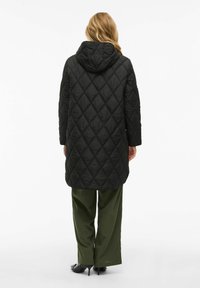 Black quilted long coat with a hood, featuring a diamond pattern, worn over green wide-leg pants and black heels.