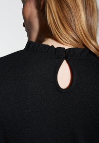Schwarzes Strickoberteil mit einem Rüschenausschnitt, das auf der Rückseite eine Schlüsselöffnungsdetails aufweist. Das Material hat einen dezenten Schimmereffekt.