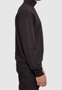 Sweat-shirt noir à col montant avec manches longues, poignets côtelés et tissu texturé. La vue de côté met en valeur sa coupe ajustée et sa finition lisse.