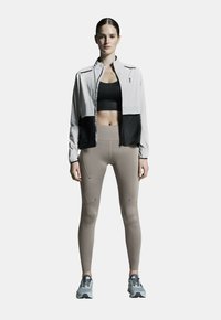 Leichte, mit Reißverschluss versehene Jacke in Grau und Schwarz, kombiniert mit beigen Leggings mit Nahtdetails. Texturiertes Material und Laufschuhe runden das Outfit ab.