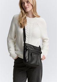 Schwarze synthetische Umhängetasche mit strukturiertem Oberflächen, verstellbarem Riemen und Front-Reißverschlusstasche, getragen über einem weißen Kabelstrickpullover.
