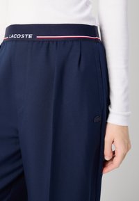 Pantaloni blu navy con vita piatta, caratterizzati da una striscia bianca e rossa, pieghe e un piccolo logo Lacoste sulla tasca laterale.