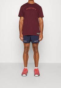 Camiseta de algodón color burdeos con el logo "PROJECT ROCK", combinada con pantalones cortos azul marino con texto blanco. Zapatillas rojas con acentos azules.