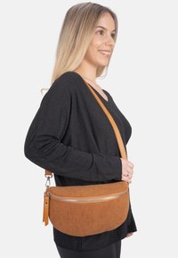 Crosbody-Tasche aus Cord in Braun mit einer runden Form, silbernem Reißverschluss und einer flachen, strukturierten Oberfläche. Verstellbarer, beigefarbener Riemen.