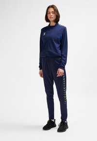 Granatowy dres wykonany z lekkiej tkaniny, składający się z kurtki na suwak oraz spodni z elastyczną talią i bocznymi wstawkami w zygzakowaty wzór. Czarne trampki.