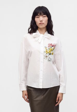 Femme portant un chemisier blanc boutonné avec col lavallière et broderie florale sur la poitrine, associé à une jupe foncée, debout sur fond blanc.