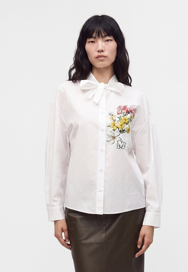Femme portant un chemisier blanc boutonné avec col lavallière et broderie florale sur la poitrine, associé à une jupe foncée, debout sur fond blanc.