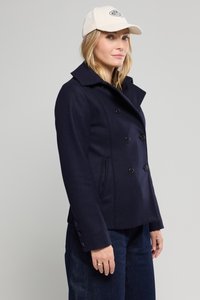 Manteau double boutonnage en mélange de laine bleu marine avec un col haut, doté de boutons noirs et de poches latérales. Associé à une casquette de baseball crème.
