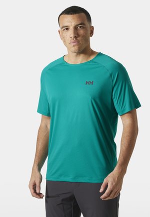 Man met kort haar en sproeten, gekleed in een groenblauw sportshirt met korte mouwen en zwarte broek, staand tegen een witte achtergrond.
